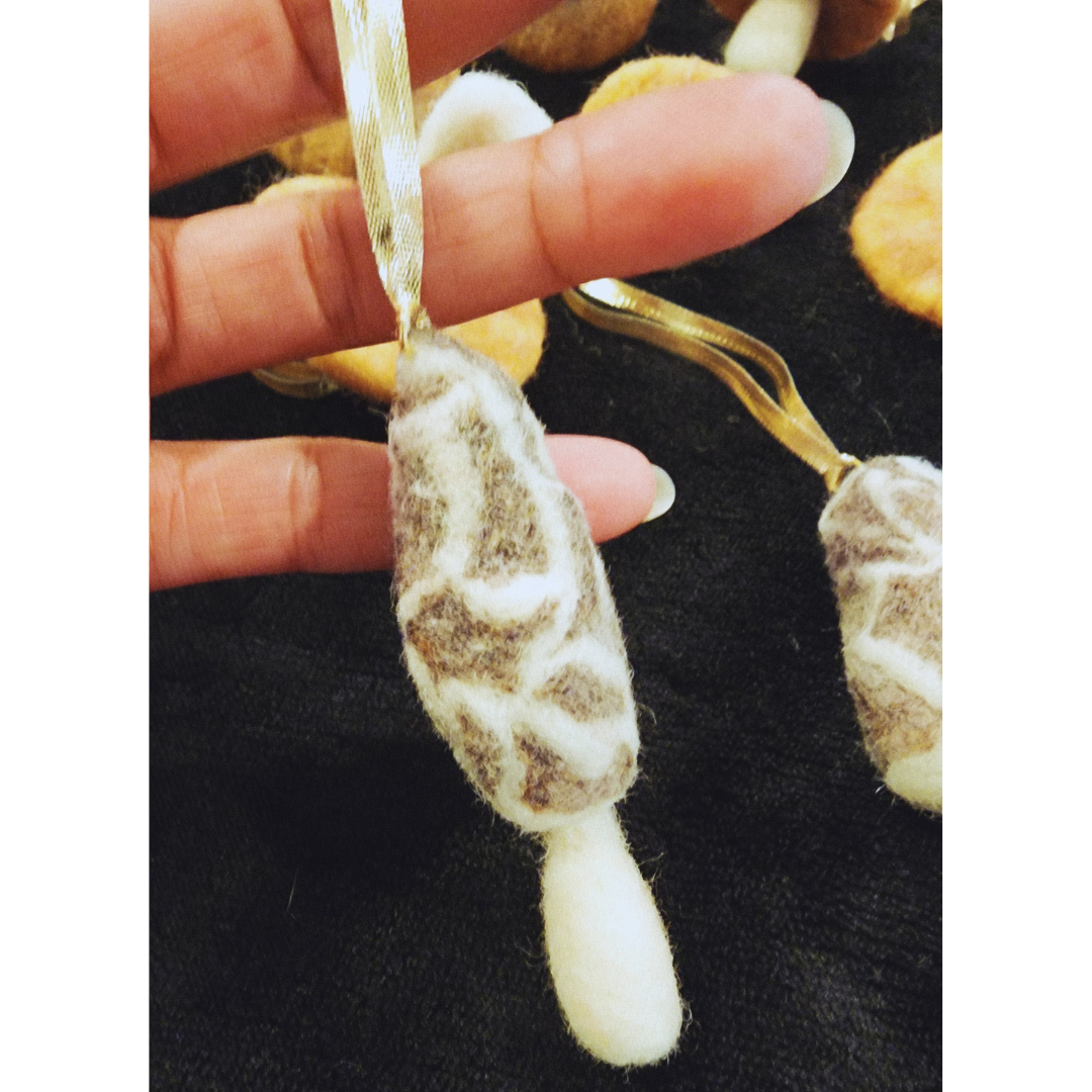 Close-up of a hand holding one long, textured needle-felted morel mushroom ornament. The morel features brown and white patterned wool and a white stem.