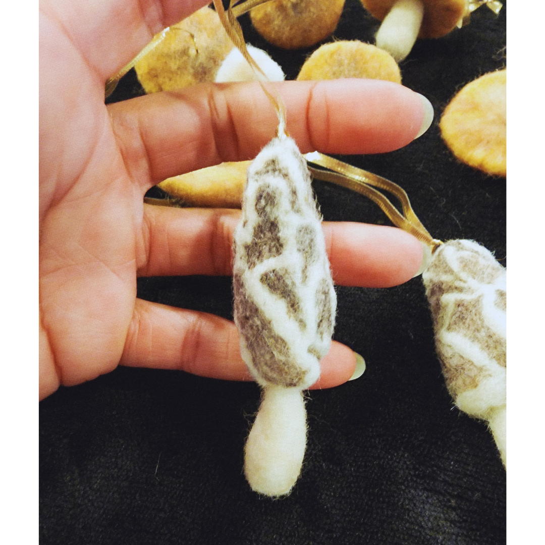 Detailed view of a hand holding the long, textured needle-felted morel mushroom ornament, highlighting its unique brown and white pattern.