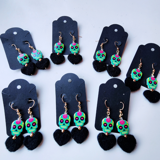 A flat lay photo of several pairs of handmade earrings on black display cards. Each earring has a colorful teal and pink sugar skull bead with a black felt heart hanging below it.