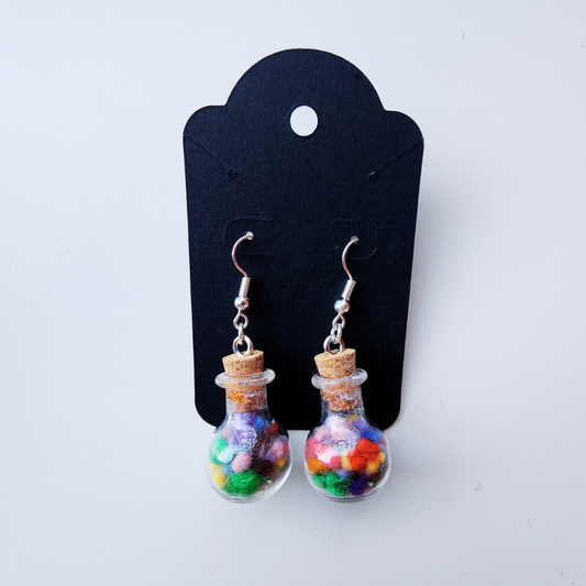 A flat lay photo showing a pair of my handmade potion bottle earrings on a black display card. The earrings are tiny glass bottles filled with colorful wool, with a cork stopper and silver findings.