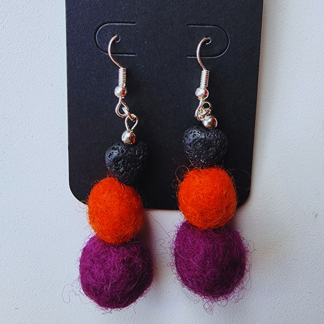 A pair of handmade earrings displayed on a black card. Each earring is made of a black heart-shaped lava bead, an orange felt ball, and a purple felt ball.