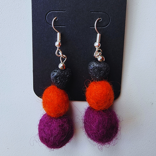 A pair of handmade earrings displayed on a black card. Each earring is made of a black heart-shaped lava bead, an orange felt ball, and a purple felt ball.