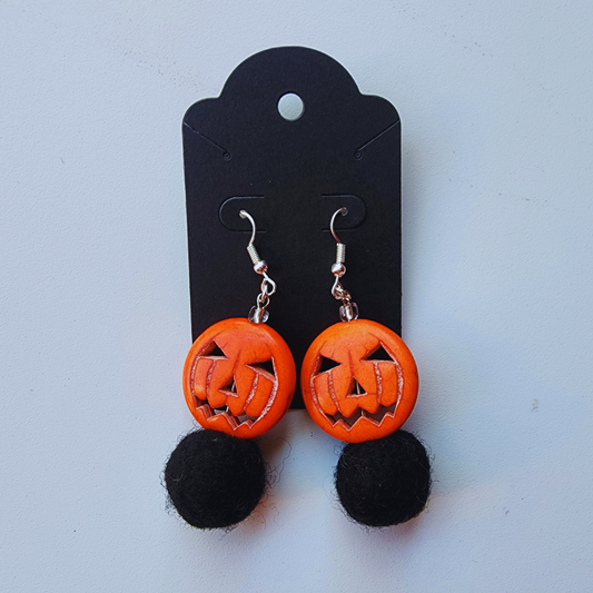 A pair of handmade earrings displayed on a black card. Each earring features a carved orange ceramic jack-o-lantern bead with a round, black felted bead hanging below it.