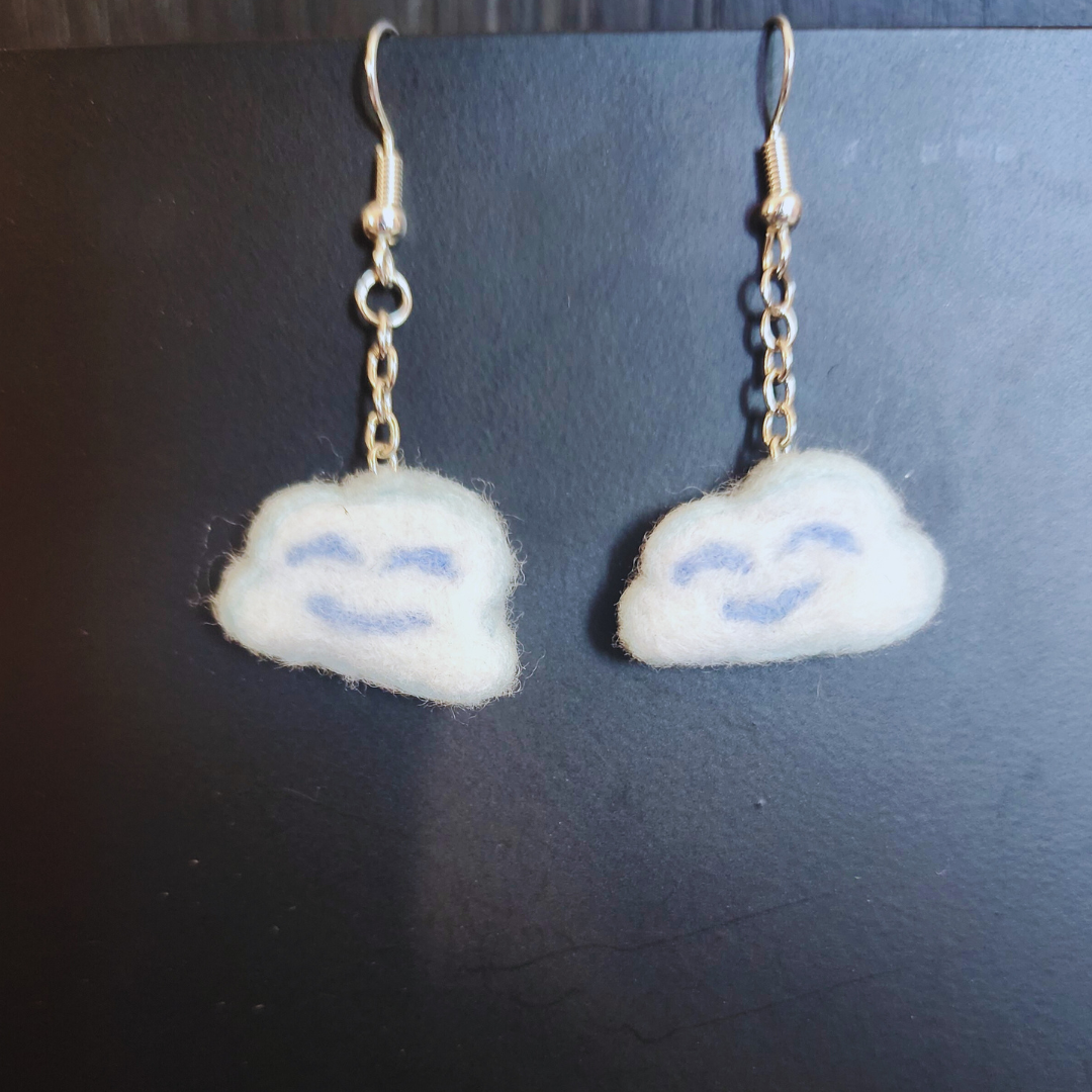 A pair of handmade light blue and white felt Kawaii Cloud Dangle Earrings with light blue smiling faces, hanging side-by-side against a dark background.
