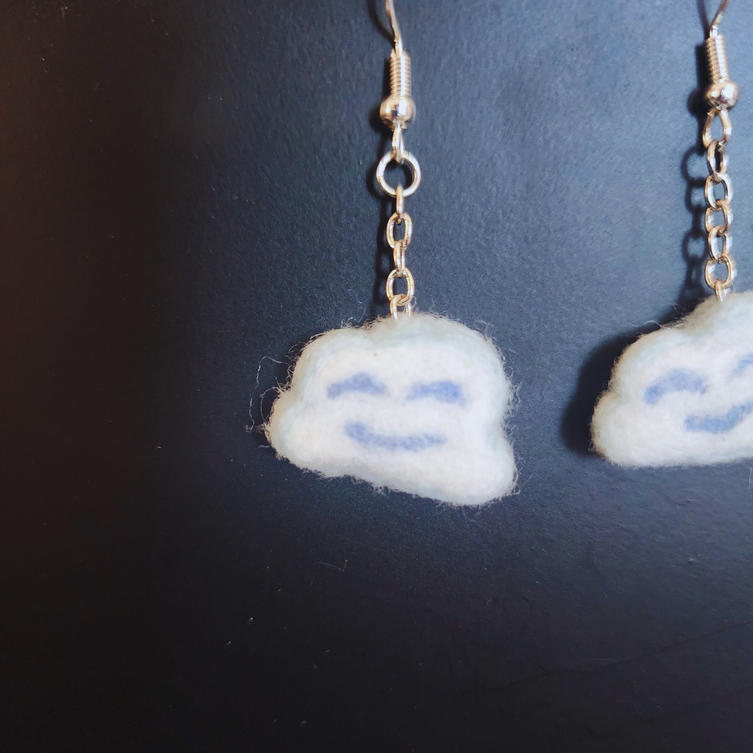A close-up of one handmade light blue and white felt Kawaii Cloud Dangle Earring with a light blue smiling face, showing the silver-toned hook and chain against a dark background.