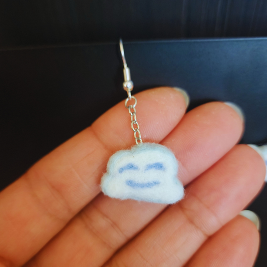 A handmade light blue and white felt Kawaii Cloud Dangle Earring with a smiling face, held between fingertips to show its size and detail.