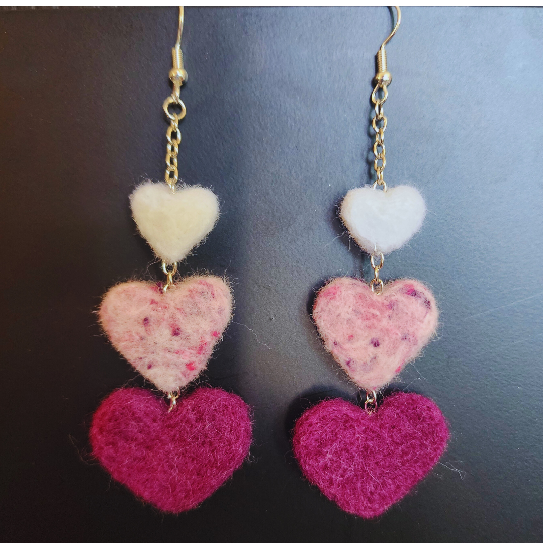 A pair of handmade needle-felted heart chain earrings, each featuring three felted hearts stacked vertically in descending shades of pink and white, with gold-toned hooks and chains, against a dark background.