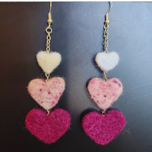A pair of handmade needle-felted heart chain earrings, each featuring three felted hearts stacked vertically in descending shades of pink and white, with gold-toned hooks and chains, against a dark background.