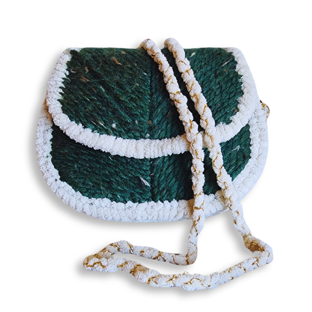 A small, handmade embroidered saddlebag in deep forest green with a white chenille border and a woven gold chain and white yarn strap draped over the front.