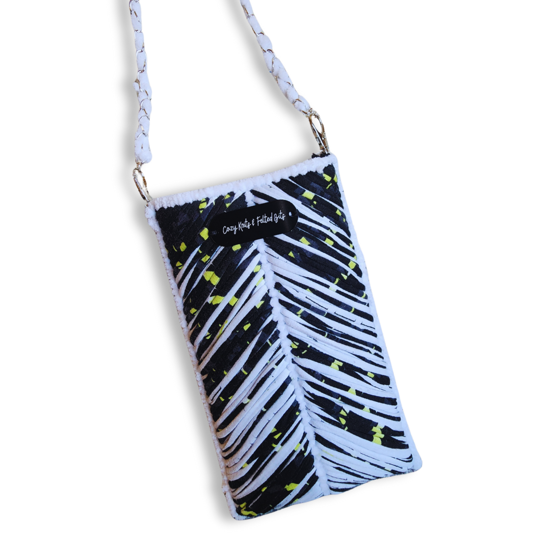 A handmade embroidered phone purse with a chevron pattern in black, white, and neon yellow, hanging from a white woven chain strap. A small "Cozy Knits & Felted Bits" tag is visible.
