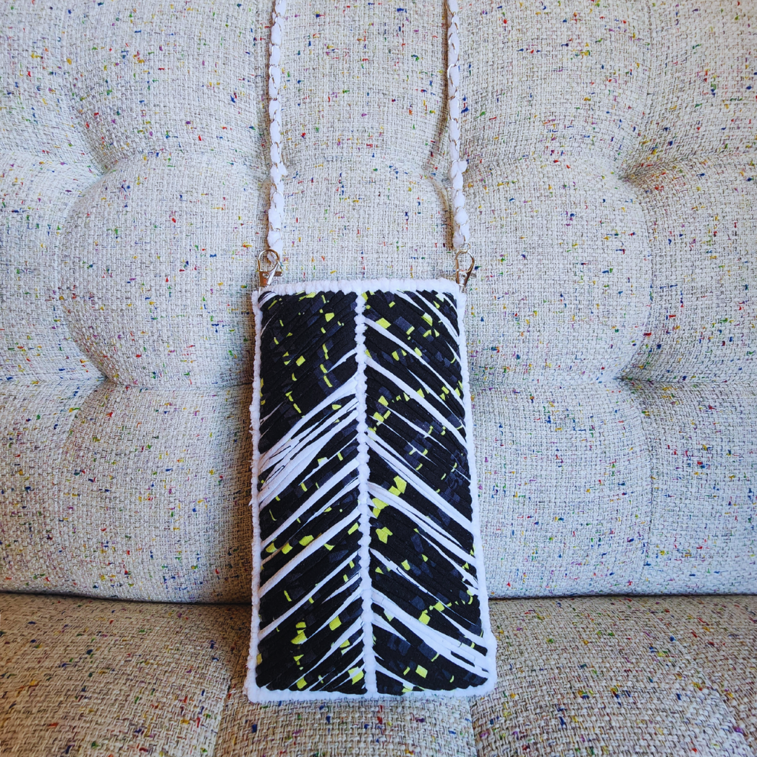 A handmade phone purse featuring a dramatic black and white chevron embroidery pattern with accents of neon yellow, hanging against a textured, multi-color tweed couch.