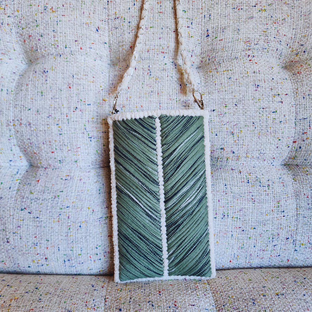 A handmade embroidered phone purse in shades of olive and dark green with a prominent chevron pattern, framed by a fuzzy white chenille edge, hanging against a textured tweed couch.