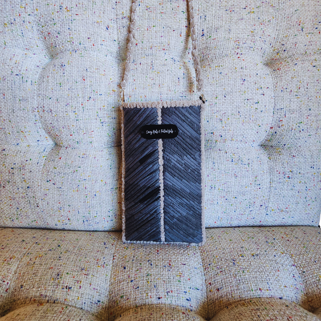 A handmade phone purse in shades of gray with a prominent chevron stitch pattern and beige chenille trim, hanging against a textured tweed couch.