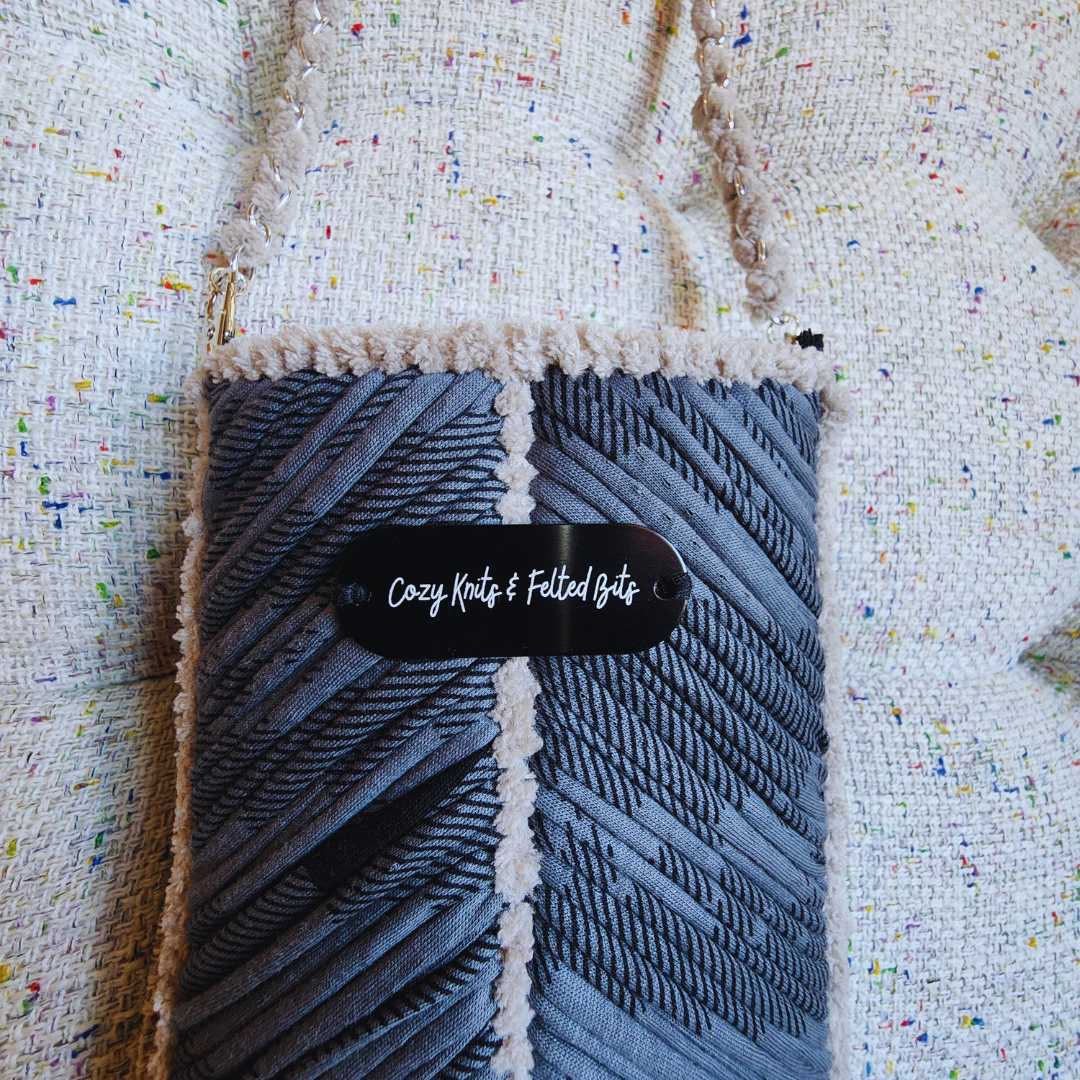 A close-up of the gray embroidered phone purse, highlighting the intricate chevron stitch, the beige chenille trim, and a black tag featuring the "Cozy Knits & Felted Bits" logo.