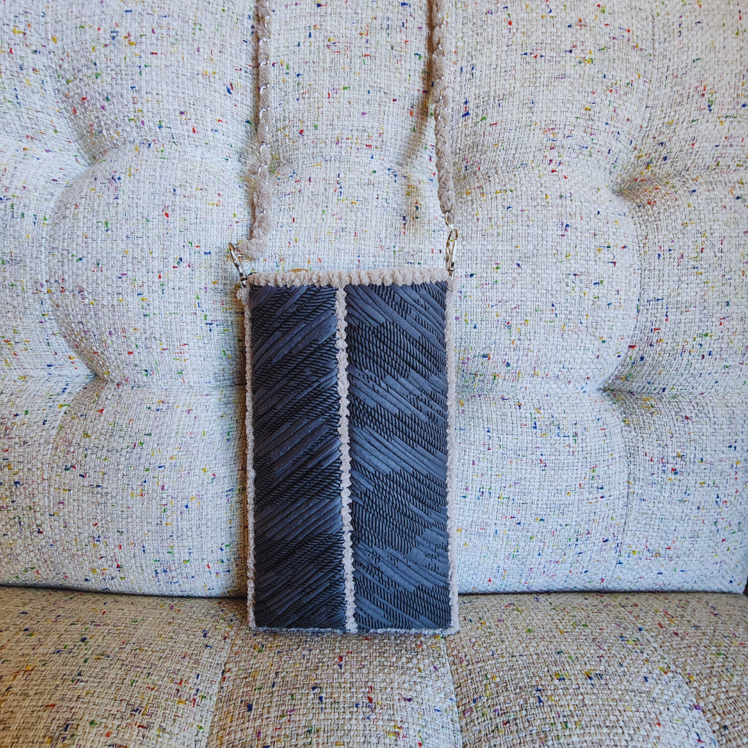 A handmade embroidered phone purse in a gray and light beige chevron pattern, hanging vertically against a textured, light tweed couch.