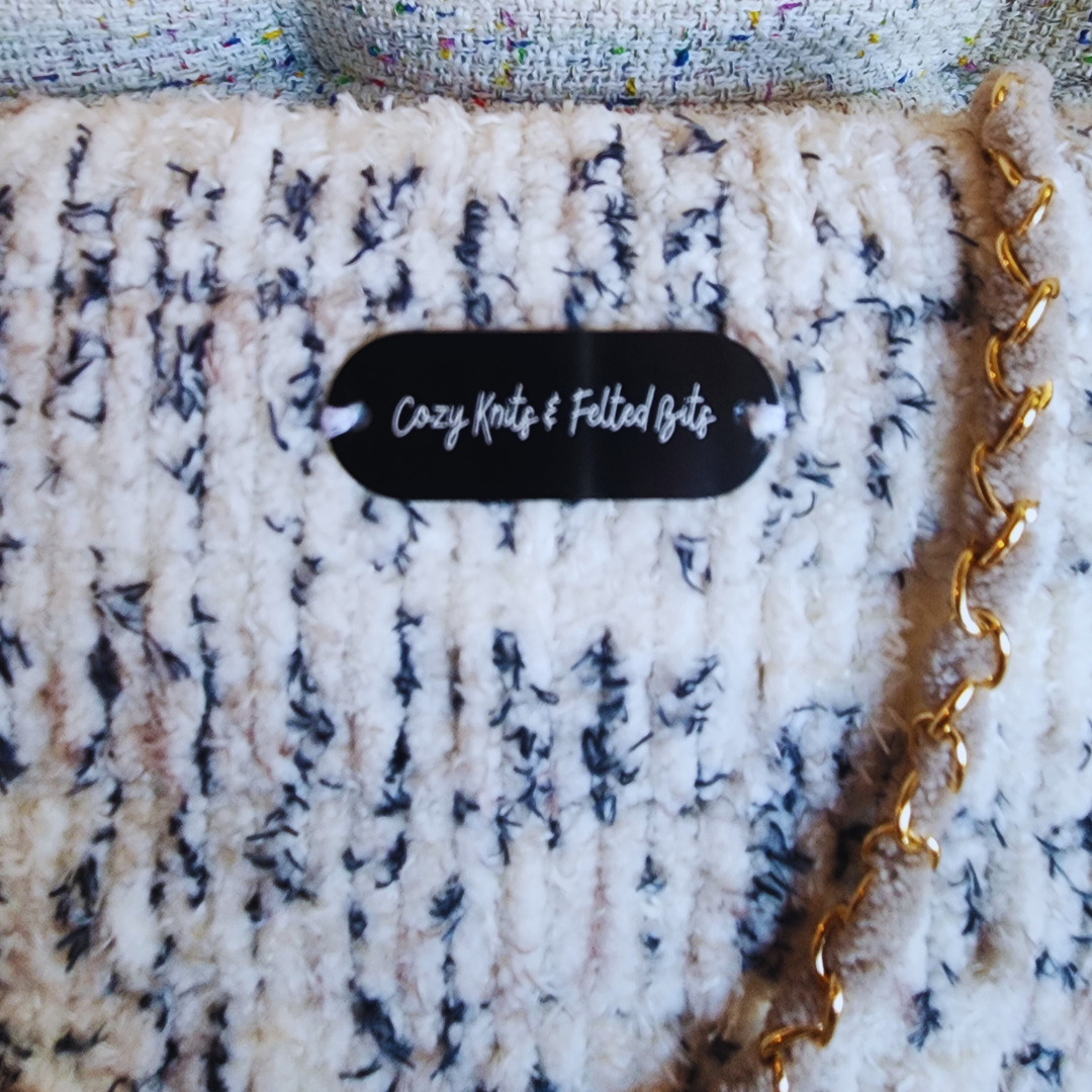 A close-up of the saddlebag's flap, showing the plush, textured white and blue/black speckled chenille yarn and the black branded "Cozy Knits & Felted Bits" tag.