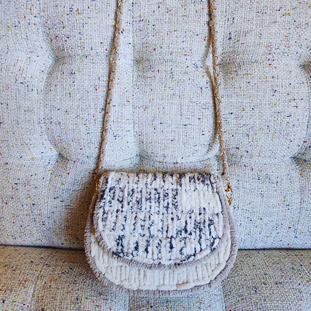 A small, handmade embroidered saddlebag in a white and blue/black speckled yarn blend, hanging against a textured, light tweed couch. The gold chain strap is visible.