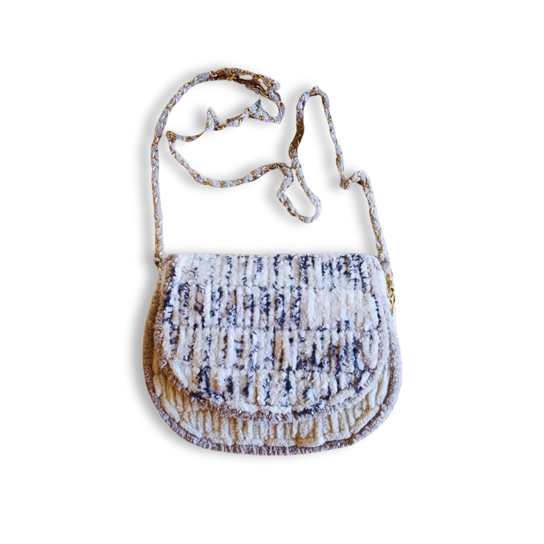A full-length view of the saddlebag with the gold and beige woven chain strap hanging down. The purse is made of textured, speckled yarn.
