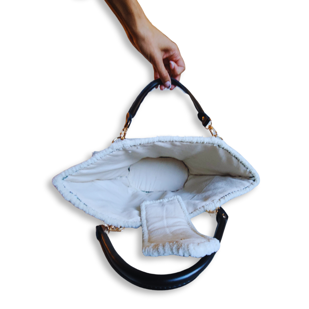 A hand holds the black leather strap of the open tote bag, revealing the wide interior, the cream lining, and the magnetic closure flap hanging down.