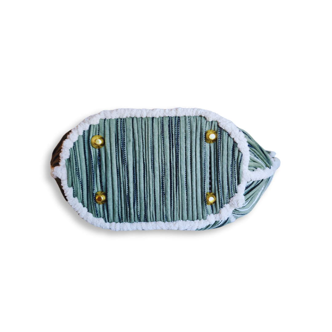 An image of the oval-shaped bottom of the tote bag, clearly showing the green and white stitching, the white chenille border, and the four protective brass purse feet.