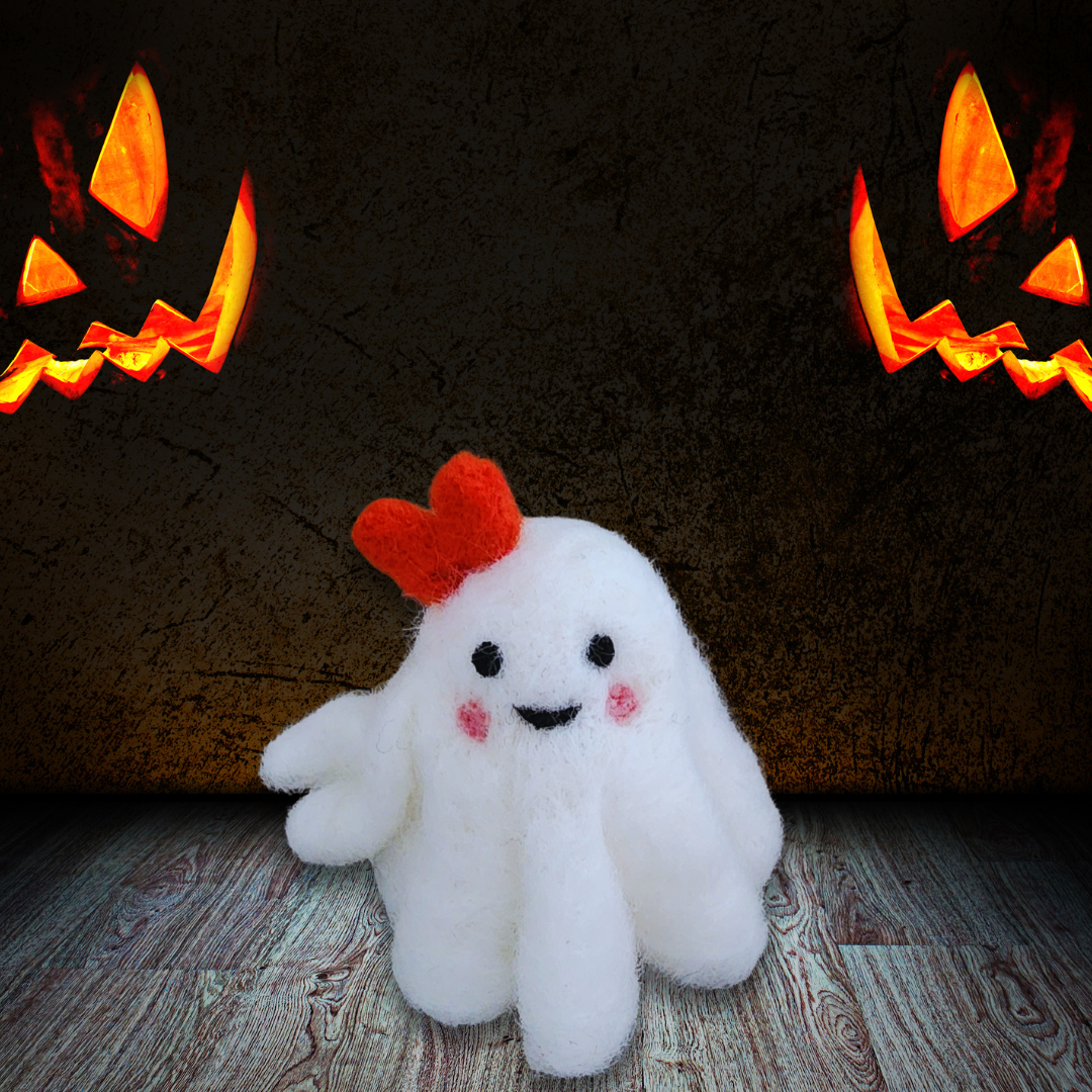 A large handmade felted ghost figurine with an orange bow, a cute smiling mouth, and pink cheeks, sitting on a rustic wooden floor with two glowing jack-o'-lanterns in the dark background.