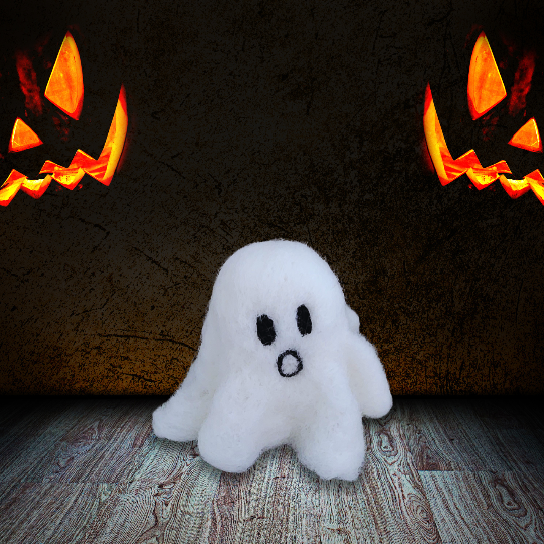  A large handmade felted ghost figurine with a surprised, open-mouthed expression and black eyes, sitting on a rustic wooden floor with two glowing jack-o'-lanterns in the dark background.