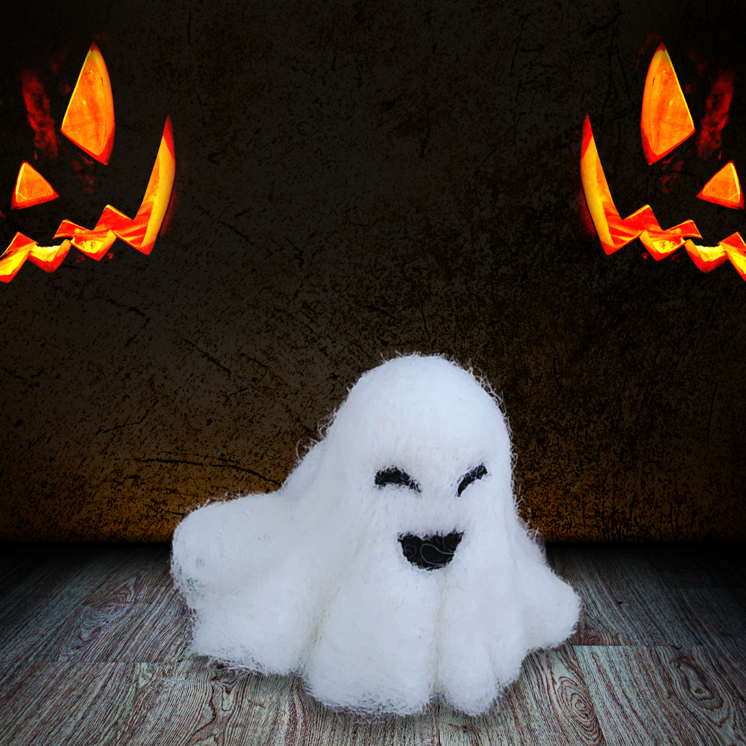 A small handmade felted ghost figurine with a wide black smiling mouth and small eyes, sitting on a rustic wooden floor with two glowing jack-o'-lanterns visible in the dark background.
