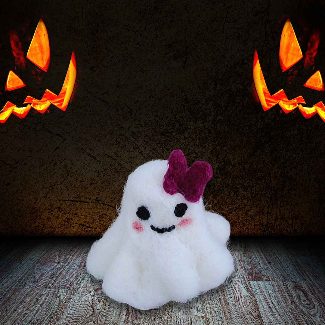 A small handmade felted ghost figurine with a purple bow, a cute smiling mouth, and pink cheeks, sitting on a rustic wooden floor with two glowing jack-o'-lanterns visible in the dark background.