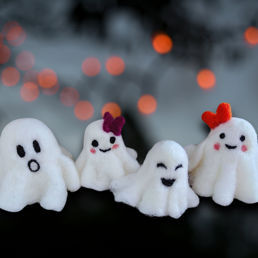 A group of four handmade felted ghost figurines with different faces and accessories (one with spooky black eyes, one with a purple bow, one with a wide smile, and one with a red heart bow), standing in a row with a blurry background of glowing orange lights.
