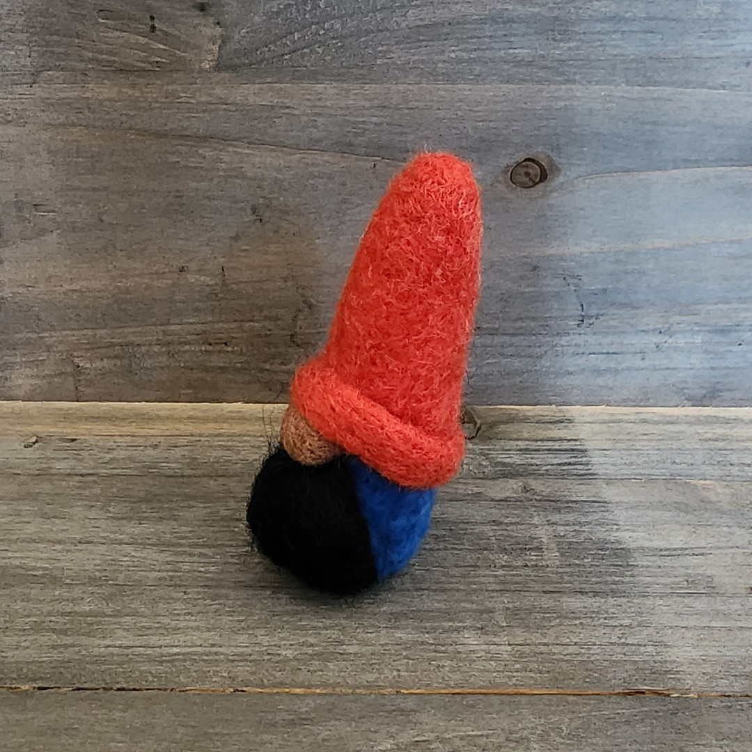A handmade needle-felted gnome with a bright red pointed hat, a brown nose, and a black beard, with a blue body partially visible, viewed from a side angle on a wooden background.