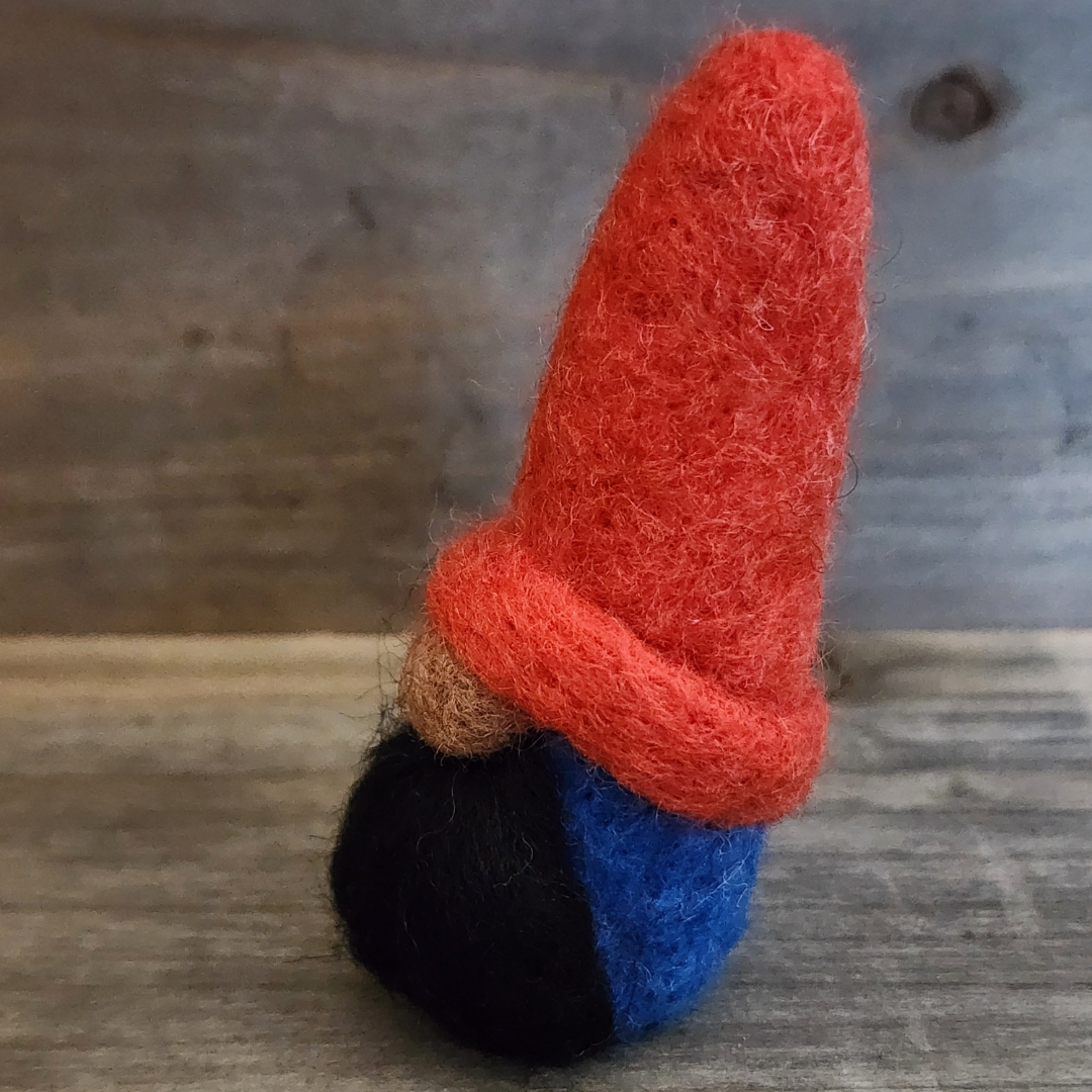 A handmade needle-felted gnome with a bright red pointed hat, a brown nose, and a black beard, with a blue body partially visible, viewed from a slightly angled perspective on a wooden background.