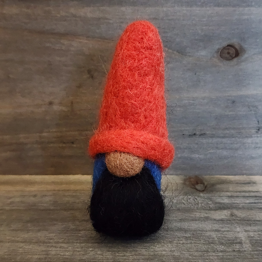 A handmade needle-felted gnome with a bright red pointed hat, a brown nose, and a full black beard, with a blue body partially visible, standing centered against a rustic wooden background.