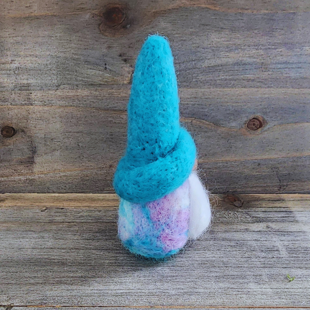 A handmade needle-felted gnome with a teal hat, a brown nose, and a fluffy white beard, with a cotton candy blended body visible below, viewed from a side angle against a rustic wooden background.