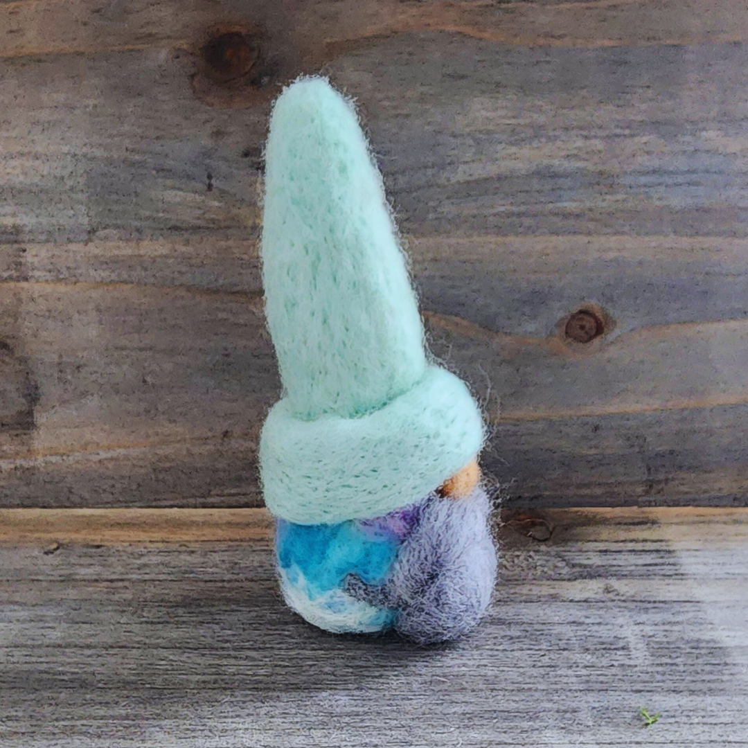 A handmade needle-felted gnome with a light mint green hat, a light-colored nose, and a fluffy gray beard, with a cotton candy blended body visible below, viewed from a side angle against a wooden background.