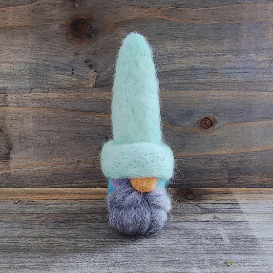 A handmade needle-felted gnome with a light mint green hat, a light-colored nose, and a fluffy gray beard, with a cotton candy blended body visible below, standing centered against a rustic wooden background.