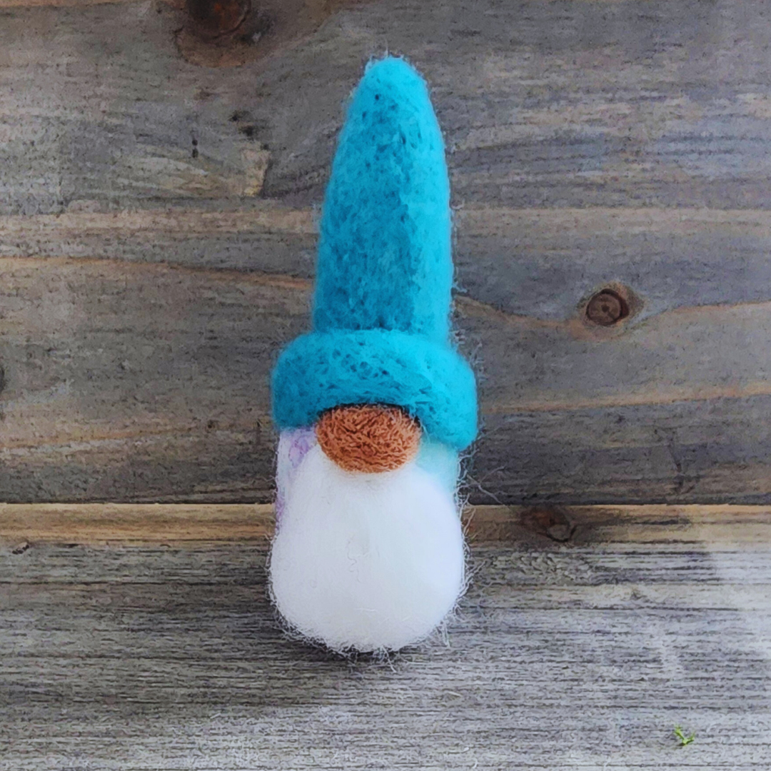 A handmade needle-felted gnome with a sky blue hat, an orange nose, and a fluffy white beard, with a cotton candy blended body visible below, standing centered against a rustic wooden background.