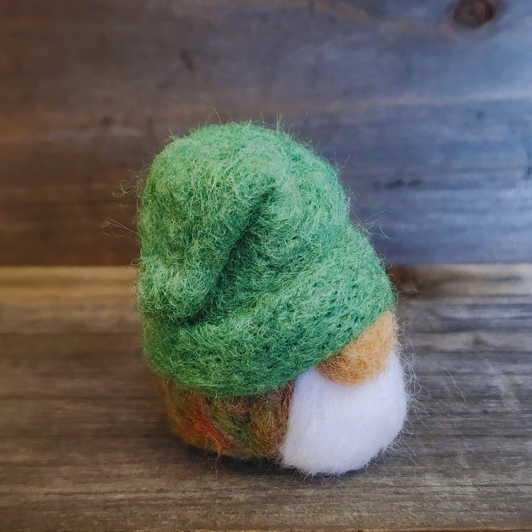 A handmade needle-felted gnome with a vibrant green pointed hat, a light-colored nose, and a fluffy white beard, standing centered against a rustic wooden background.