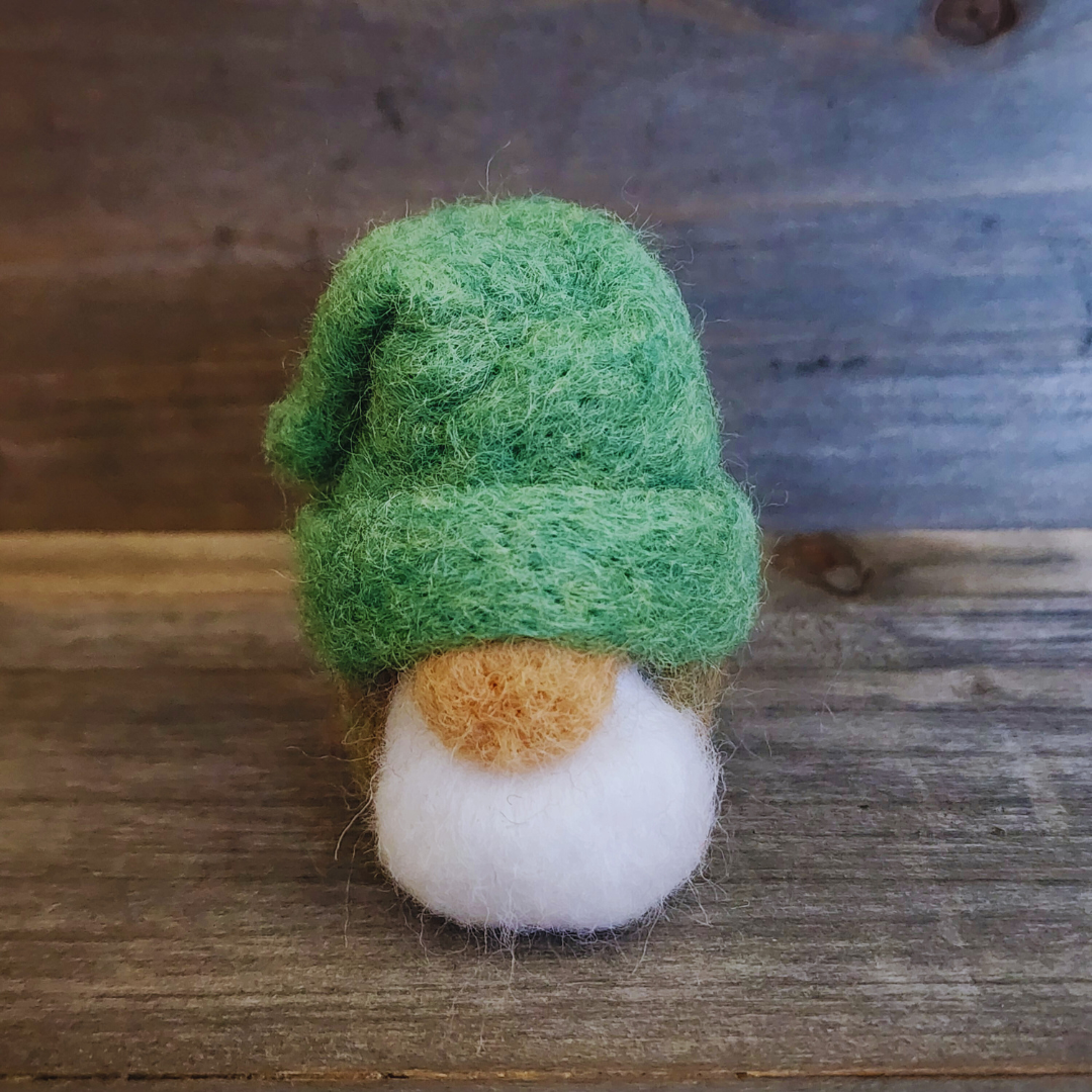A handmade needle-felted gnome with a vibrant green hat, a light-colored nose, and a fluffy white beard, standing centered against a rustic wooden background.