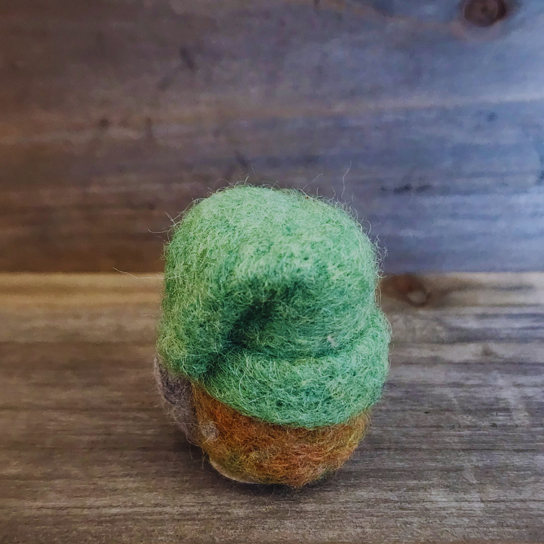 A handmade needle-felted gnome with a vibrant green hat and a light-colored nose, viewed from slightly above, showing its rounded shape, against a rustic wooden background.