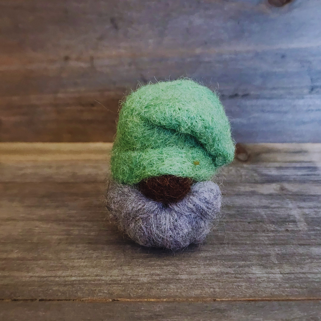 A handmade needle-felted gnome with a vibrant green hat, a small dark nose, and a greyish-purple beard, standing centered against a rustic wooden background.