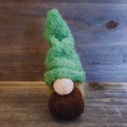 A handmade needle-felted gnome with a vibrant green pointed hat, a light-colored nose, and a brown beard, standing centered against a rustic wooden background.