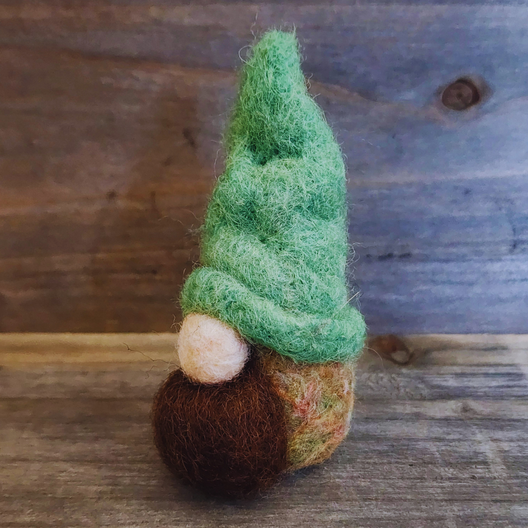 A handmade needle-felted gnome with a vibrant green pointed hat, a light-colored nose, and a brown beard, viewed from a slight side angle against a rustic wooden background.