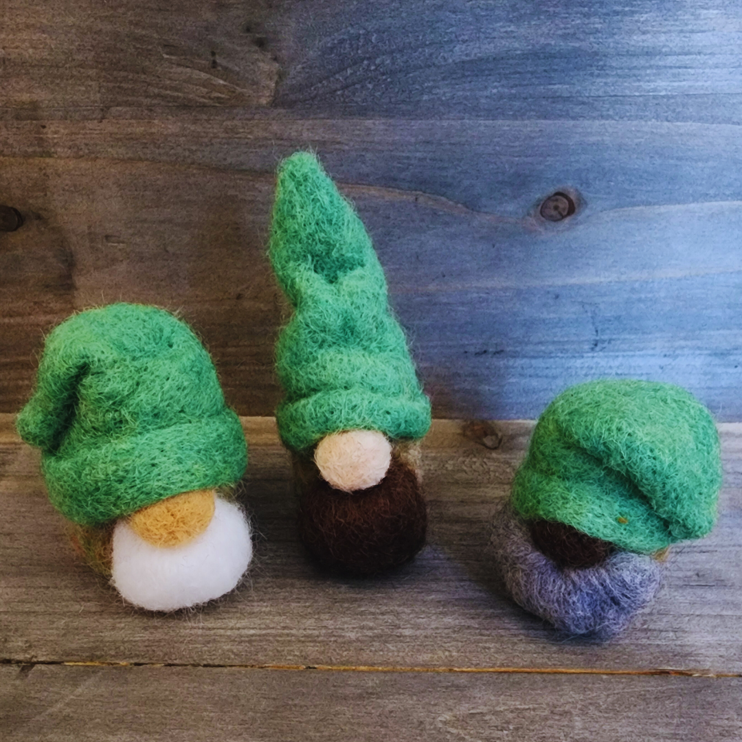 A group of three handmade needle-felted gnomes with varying green hats, beards, and body colors, standing together on a rustic wooden surface.