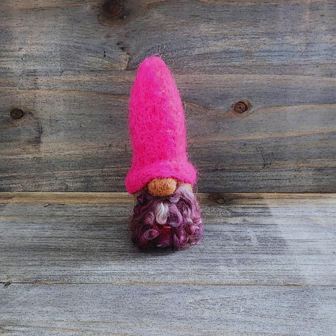 A handmade needle-felted gnome with a hot pink pointed hat, a mixed purple curly beard, and a light-colored nose, standing centered against a rustic wooden background.
