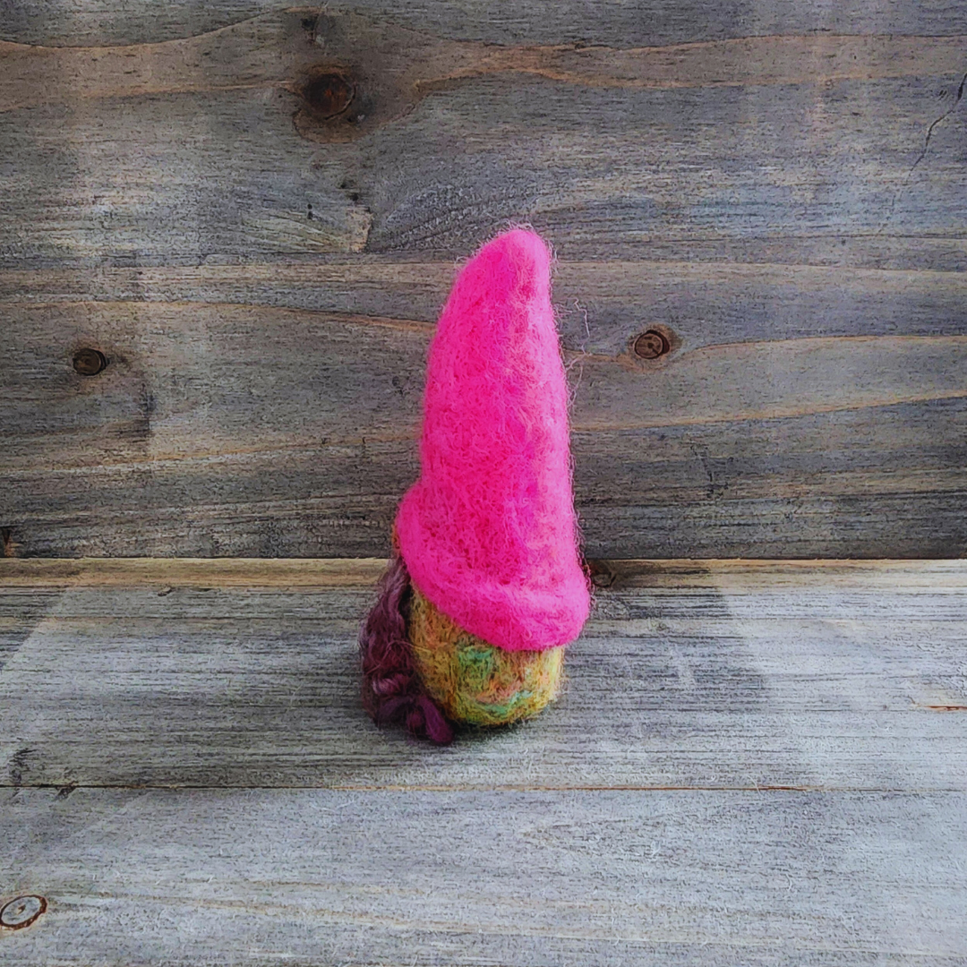 A handmade needle-felted gnome with a hot pink pointed hat and a blended green merino wool body with a hint of brown beard, viewed from a side angle against a wooden background.