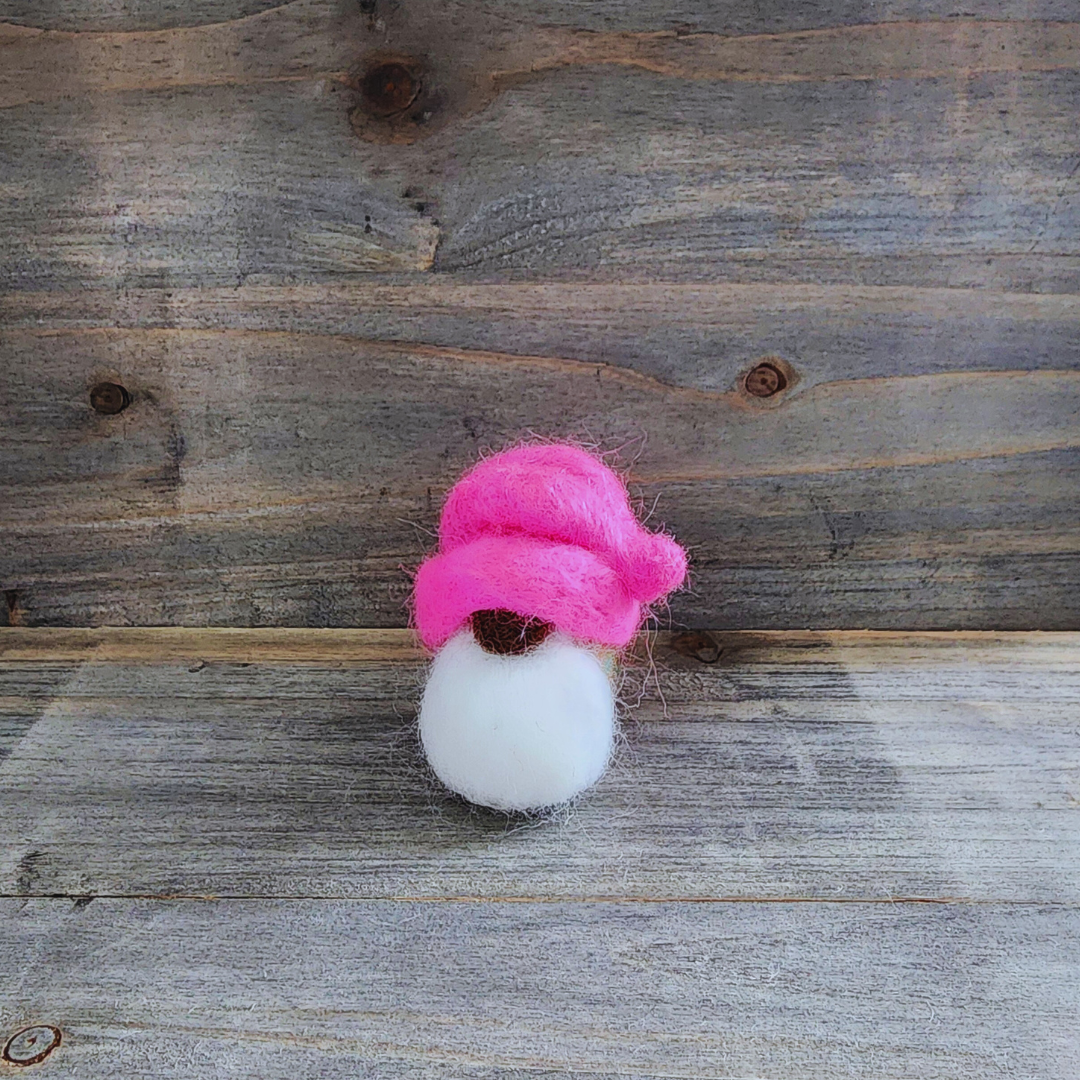 A small handmade needle-felted gnome with a hot pink hat, a brown nose, and a fluffy white beard, standing centered against a rustic wooden background.