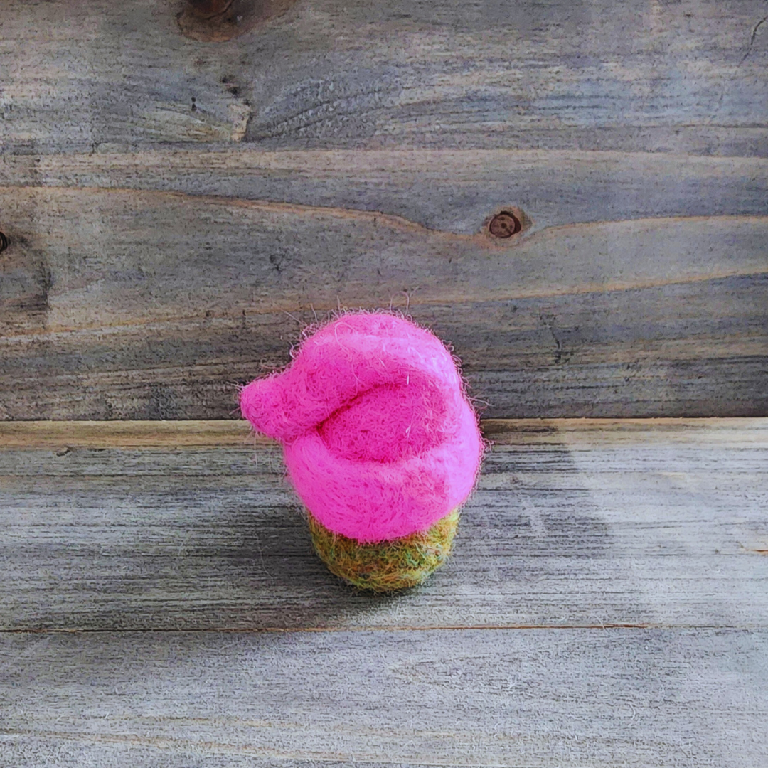 An overhead view of a handmade needle-felted gnome with a hot pink hat, showing the top of its blended green merino wool body and the hat's rounded shape, on a wooden background.