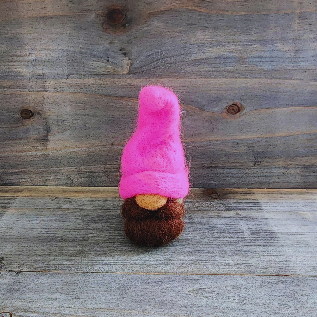 A handmade needle-felted gnome with a hot pink pointed hat, a brown beard, and a light-colored nose, standing centered against a rustic wooden background.