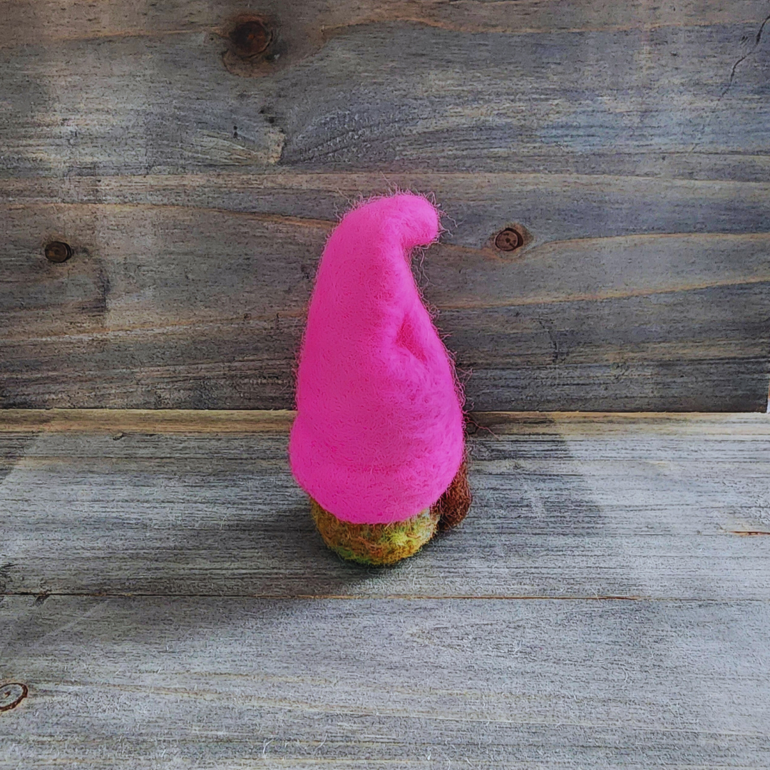 A handmade needle-felted gnome with a hot pink pointed hat and a blended green merino wool body with a hint of brown beard, viewed from a side angle against a rustic wooden background.