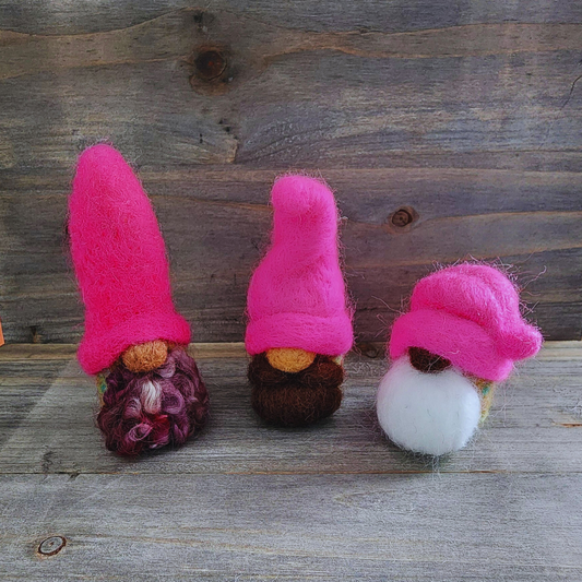 A group of three handmade needle-felted gnomes with hot pink hats and varying beard styles (curly mixed purple, solid brown, and white), standing on a wooden surface.
